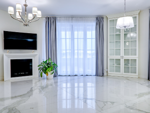 Bright living room with white marble floors, a fireplace with a wall-mounted TV, a green potted plant, and a glass-front cabinet by large windows with sheer curtains.